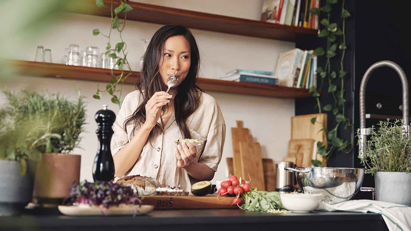 Une femme est dans la cuisine en train de cuisiner et lèche une cuillère. 