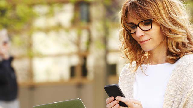 Une femme à lunettes se tient à l'extérieur avec son smartphone.